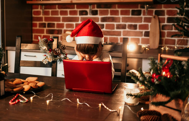 un niño caucásico en un suéter blanco y un sombrero rojo de Navidad está sentado en una mesa en la cocina y mirando un ordenador portátil - Foto, Imagen
