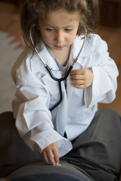 Una niña con un abrigo médico examina a su abuelo con un estetoscopio. juegos infantiles - Foto, imagen
