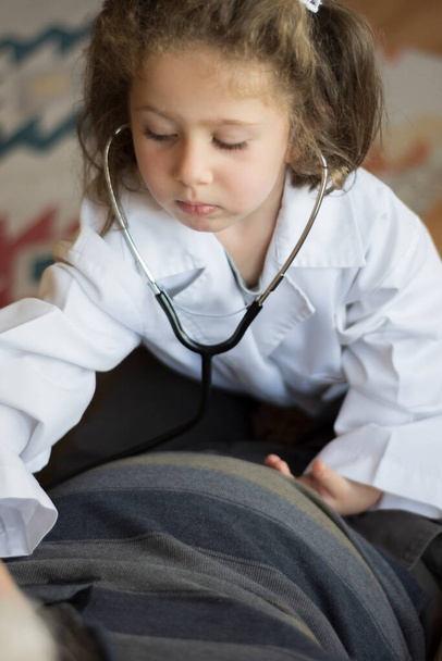 Une petite fille vêtue d'un manteau de médecin examine son grand-père avec un stéthoscope. jeux pour enfants - Photo, image