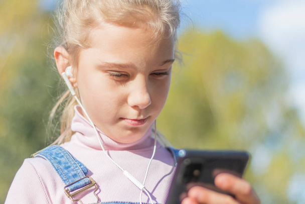 Portrait of a young beautiful Caucasian happy girl about 10 years old with long blonde hair and headphones in her ears. Cute white girl walking in the park, close-up - Foto, imagen