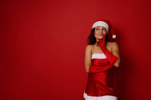 Weihnachts- und Neujahrskonzept für Werbung. Pensive geheimnisvolle schöne Frau im Weihnachtsmann-Faschingskostüm hält ihren Finger an ihre Lippen und blickt nachdenklich zu einer Werbefläche auf rotem Hintergrund auf. - Foto, Bild