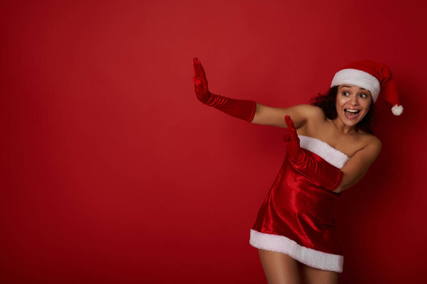 Mulher hispânica incrível vestida com roupas de Papai Noel detém um espaço de cópia em um fundo vermelho e se inclina para longe dele. Estúdio conceitual filmado para anúncio de Natal e Ano Novo com emoções humanas. - Foto, Imagem