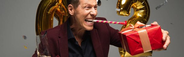 Excited man with party horn, champagne and gift near balloons on grey background, banner  - Fotografie, Obrázek