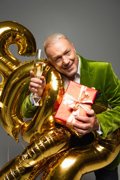 Mature man holding gift and glass of champagne near balloons isolated on grey  - Foto, Bild