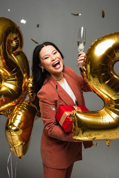 Excited asian woman holding glass of champagne and present near confetti and balloons on grey background  - Foto, imagen