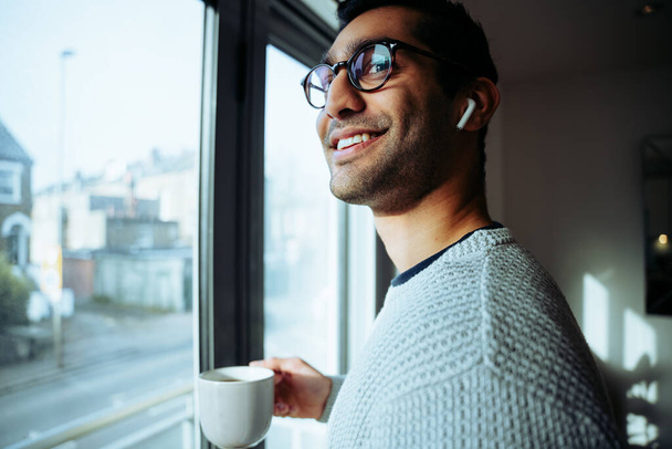 Uomo d'affari di razza mista sorridente che lavora da casa gustando caffè mattutino caldo ascoltando musica con auricolari - Foto, immagini