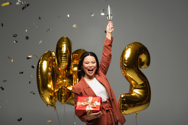 Excited asian woman holding present and champagne near balloons and confetti on grey background  - Photo, Image