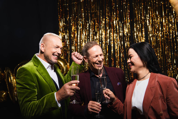 Cheerful interracial friends holding glasses of champagne near tinsel on black background  - Fotografie, Obrázek