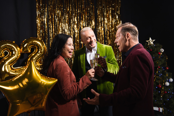 Excited man holding champagne and looking at interracial friends near christmas tree and balloons on black background  - 写真・画像
