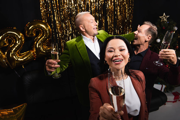 Vista ad alto angolo di donna asiatica con un bicchiere di champagne vicino agli amici e decorazioni festive su sfondo nero  - Foto, immagini