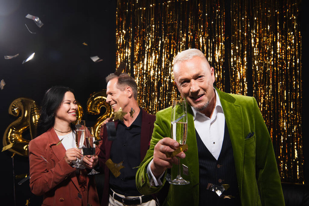 Smiling mature man holding champagne near interracial friends, confetti and tinsel on black background  - Photo, Image
