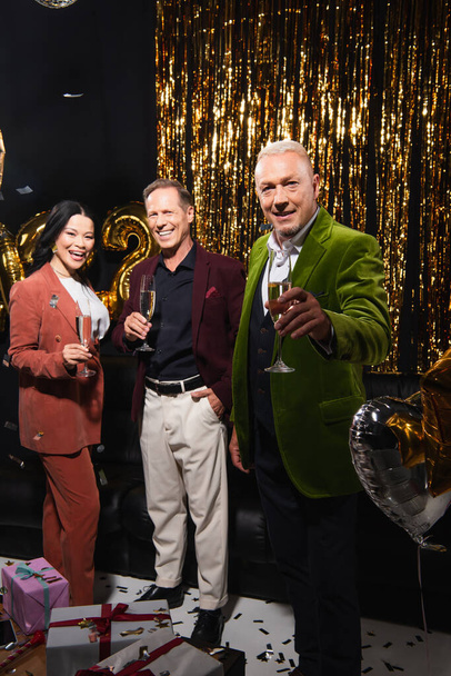 Positive mature man holding champagne near multiethnic friends, balloons and gifts on black background  - Photo, Image