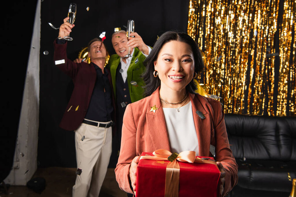 Positive asiatische Frau hält Geschenk in der Nähe verschwommener Freunde mit Champagner während der Neujahrsparty auf schwarzem Hintergrund  - Foto, Bild