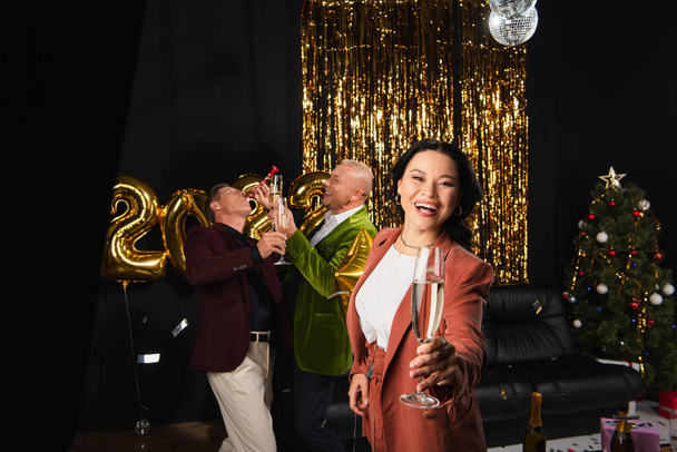 Positive asian woman holding champagne near friends with party horn and festive decor on black background  - Photo, Image