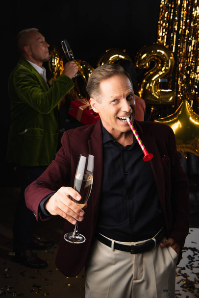 Улыбающийся взрослый мужчина с рогом для вечеринок и шампанским, смотрящий в камеру рядом с размытым другом и праздничным декором на черном фоне  - Фото, изображение