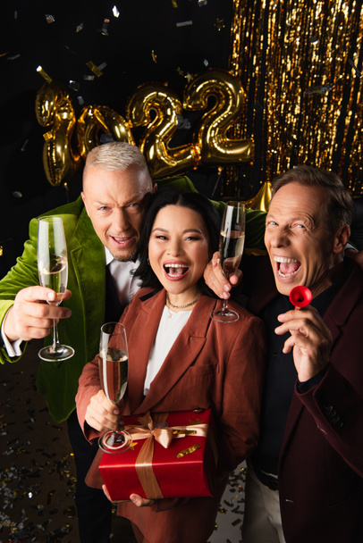 Высокоугольный вид возбужденных межрасовых друзей с шампанским и подарком, смотрящих в камеру во время новогодней вечеринки на черном фоне  - Фото, изображение