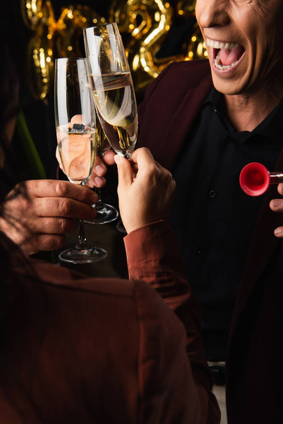 Cropped view of excited man with party horn clinking champagne with friends on black background  - 写真・画像