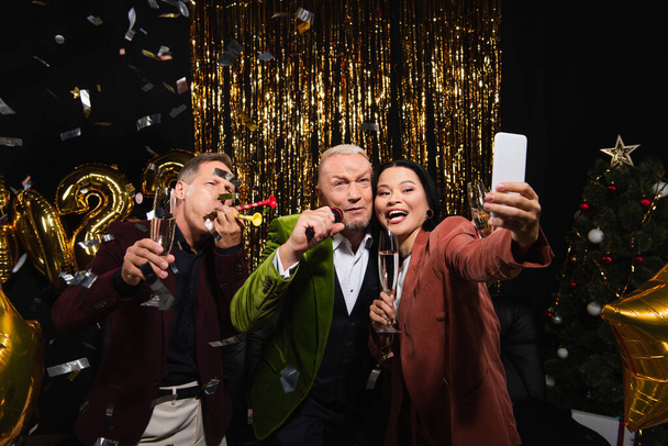 Souriant asiatique femme avec champagne prise selfie avec des amis avec microphone et cornes de fête sur fond noir  - Photo, image