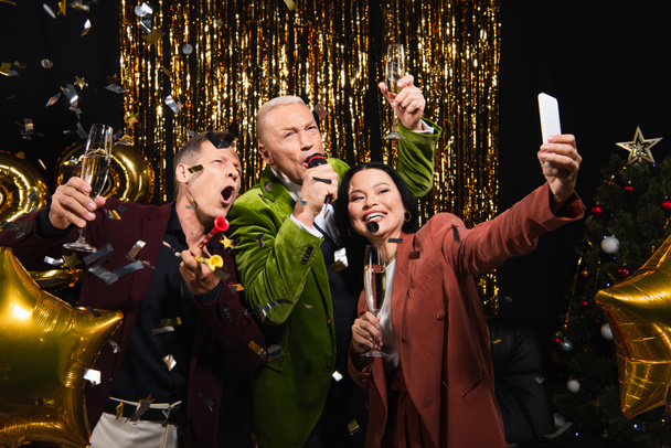 Podekscytowani międzyrasowi przyjaciele robiący selfie i śpiewający karaoke podczas noworocznej imprezy na czarnym tle  - Zdjęcie, obraz
