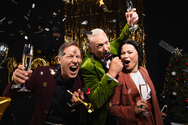 Возбужденные многонациональные друзья с шампанским поют караоке под конфетти во время вечеринки на черном фоне  - Фото, изображение