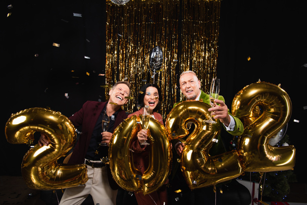Happy multiethnic mature friends holding champagne near balloons in shape of 2022 on black background  - 写真・画像