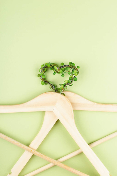 Bewust verbruik slow fashion concept. Hart van kleding hangers verstrengeld met plant op groene achtergrond.  - Foto, afbeelding