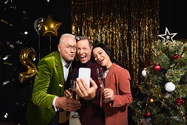 Excited multiethnic friends with champagne taking selfie on smartphone during new year party on black background  - Photo, Image