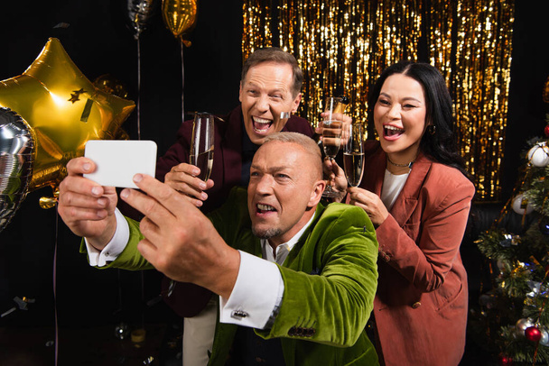 Межрасовые друзья с шампанским делают селфи на смартфоне рядом с праздничным декором на черном фоне  - Фото, изображение