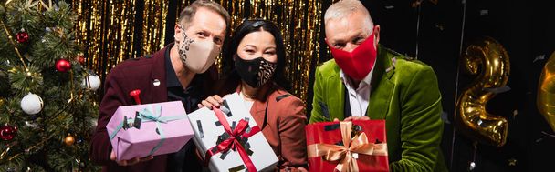 interraciale volwassen vrienden in beschermende maskers holing cadeautjes in de buurt van kerstboom op zwarte achtergrond, banner  - Foto, afbeelding