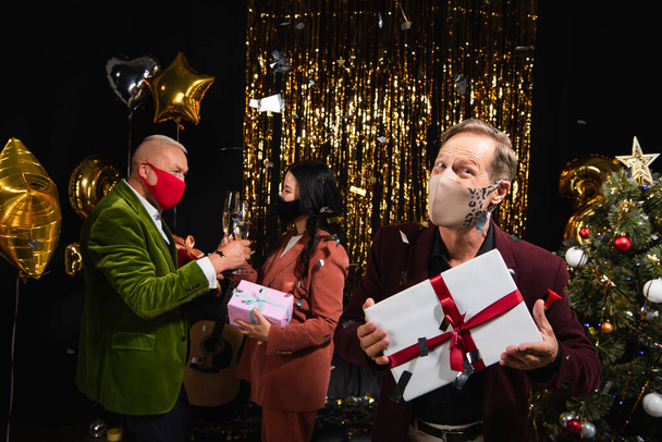 Mature man in protective mask holding present near interracial friends with champagne during new year party on black background  - Photo, Image