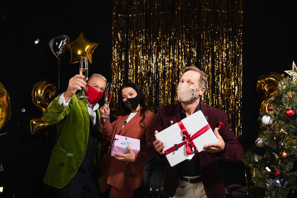 Mature interracial amis dans des masques de protection tenant du champagne et des cadeaux près de l'arbre de Noël et des ballons sur fond noir  - Photo, image