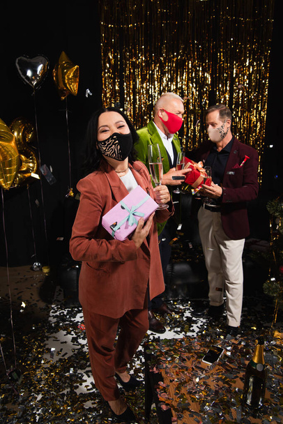 Asian woman in protective mask holding champagne and present near friends during new year party on black background  - Photo, Image