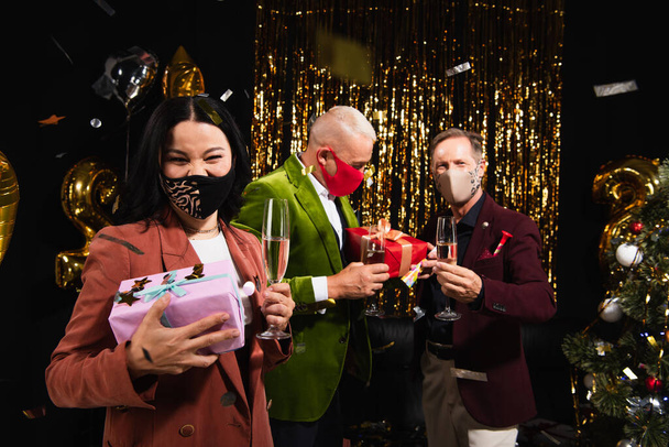 Mujer asiática en máscara protectora sosteniendo champán y regalo cerca de amigos, confeti y árbol de Navidad sobre fondo negro  - Foto, Imagen