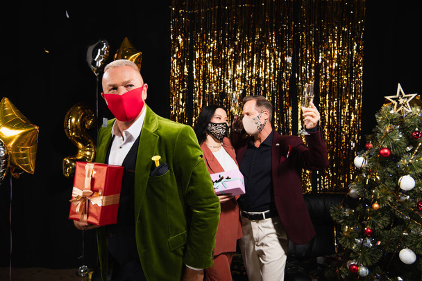 Mature man in protective mask holding gift near multiethnic friends with champagne and christmas tree on black background  - Foto, Imagem