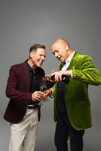 Reifer Mann gießt Champagner in Gläser neben Freund in formeller Kleidung isoliert auf grau  - Foto, Bild