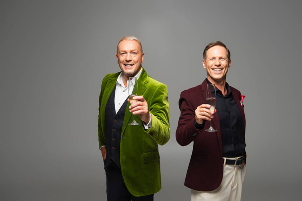 Uśmiechnięci dojrzali mężczyźni w formalnym ubraniu trzymający szampana na szaro  - Zdjęcie, obraz