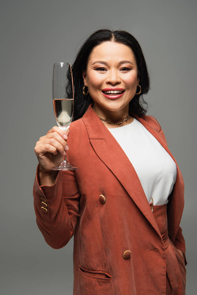 Mature asian woman holding glass of champagne and smiling at camera isolated on grey  - Foto, Imagem