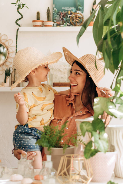 A happy young woman holds her daughter in her arms, they laugh and try on straw hats.Bright cozy home interior with indoor plants.Family, vacation, summer concept. - Foto, imagen