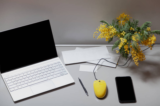 open laptop with a yellow mouse stands on a gray table. Nearby is a mobile phone business card and a bouquet of yellow mimosa. View from abov - Photo, image