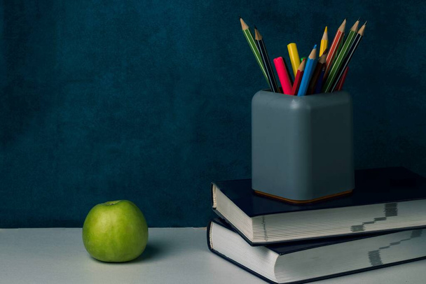 Μια στοίβα βιβλία, ένα πράσινο μήλο και ένα ποτήρι χαρτικά με πολύχρωμα μολύβια στο τραπέζι. Επιστροφή στο σχολείο έννοια - Φωτογραφία, εικόνα