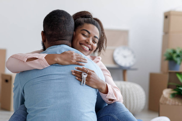 Lächelnde afrikanisch-amerikanische Ehefrau umarmt Ehemann in neuer Wohnung, Dame hält Schlüssel im Zimmer mit Schachteln - Foto, Bild