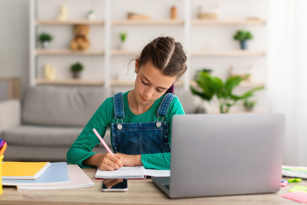 Tyttö istuu pöydän ääressä, käyttää kannettavaa tietokonetta, kirjoittaa hänen oppikirjassa - Valokuva, kuva