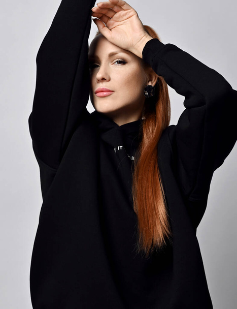 Portrait of gorgeous sexy young woman with healthy red hair in black sportswear holding arms up and hand at forehead - Photo, image