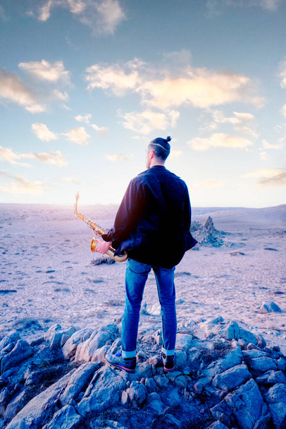 латинский молодой человек, держащий саксофон и смотрящий на горизонт пустыни - Фото, изображение