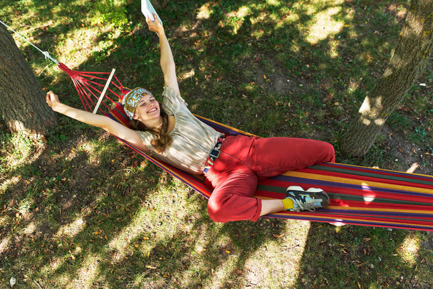 Horizontal bottom view of a young woman relaxing in hammock in the park. Pretty female resting on hammock hanging between trees in a forest. - Foto, Imagem