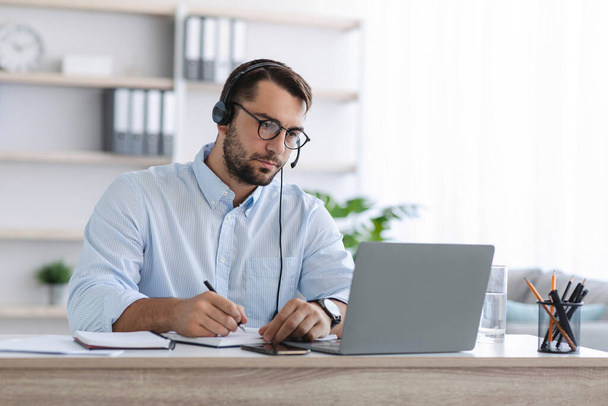 Homme européen adulte occupé manager avec barbe dans les lunettes et les écouteurs travaillent à l'ordinateur portable dans le bureau - Photo, image