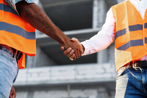 Two men engineers in workwear shaking hands against construction site. - Φωτογραφία, εικόνα