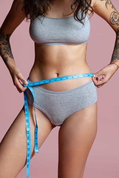 Cropped portrait of beautiful, natural female body in gray inner wear, measuring waist isolated over pink background. Body positivity - Foto, afbeelding