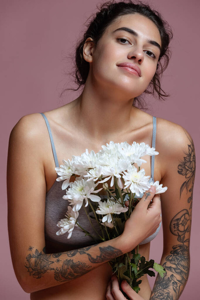 Cropped portrait of beautiful young woman in gray inner wear holding flowers isolated over pink background - Fotó, kép