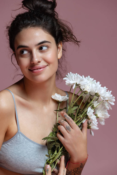 Cropped portrait of beautiful young woman in gray inner wear holding flowers isolated over pink background. Tenderness - 写真・画像
